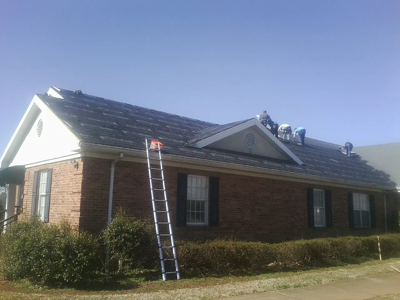 Crew tearing shingles off a brick ranch roof with a ladder set — Metal Roof Repair work by our crew in Genesee