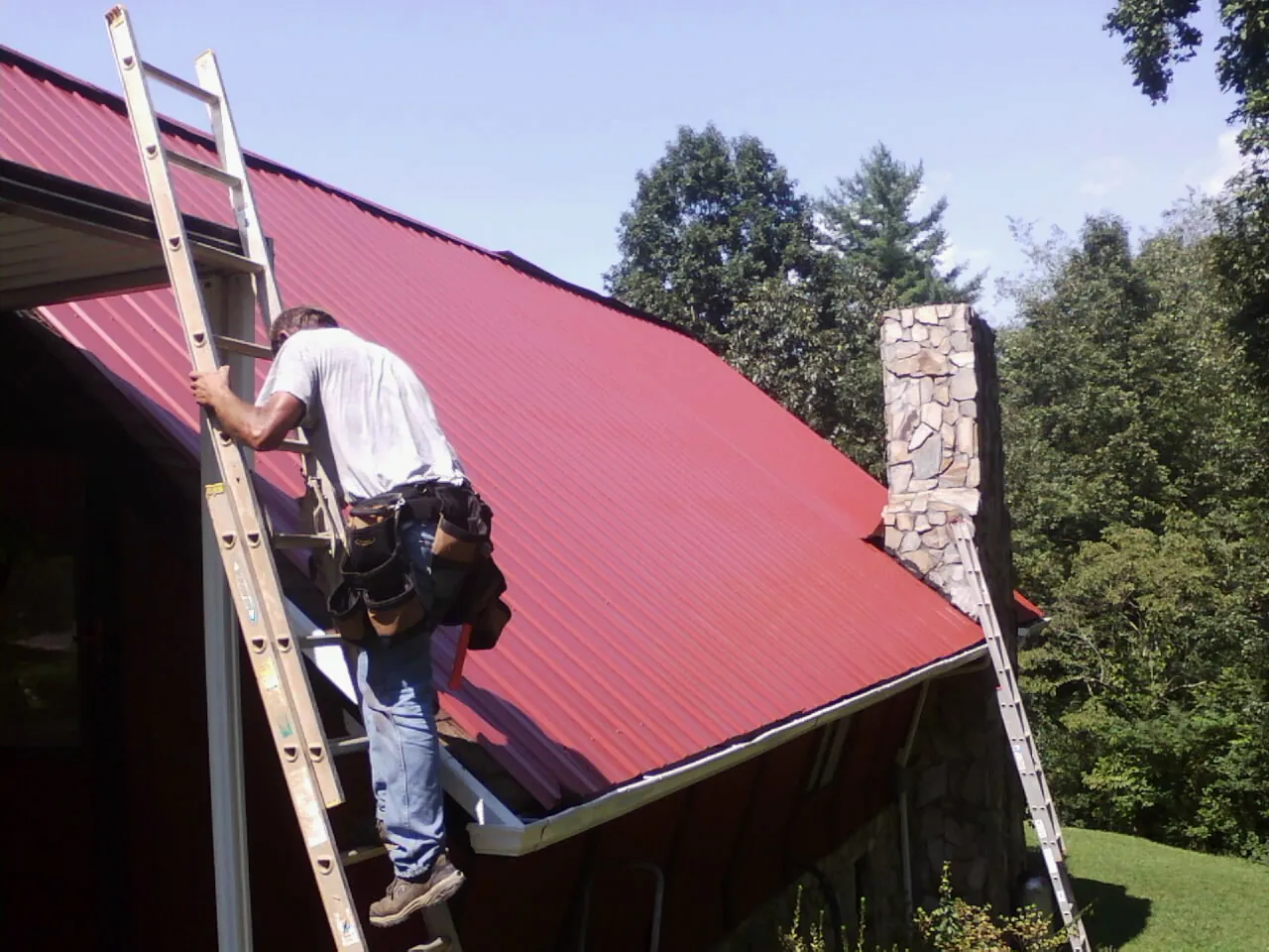 Roofer on a ladder beside a red metal roof and stone chimney — Soffit & Fascia Repair by Roofing Brothers in Genesee